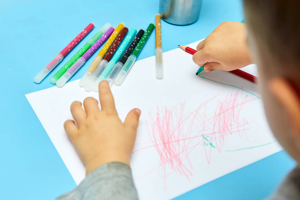 Sarı saçlı sevimli küçük çocuk evde renkli kalemler çiziyor. Mavi masaya çizim yapıyor. Beyaz kağıda resim çizen çocuğun ellerini kapat - Fotoğraf, Görsel