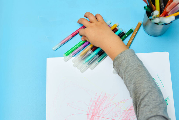 Χαριτωμένο αγοράκι με ξανθά μαλλιά ζωγραφίζει χρωματιστά μολύβια στο σπίτι. Ζωγραφίζει στο μπλε τραπέζι. Κλείσιμο των χεριών του παιδιού σε λευκό χαρτί - Φωτογραφία, εικόνα