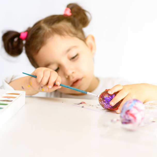 Beautiful small girl painted Easter eggs, time at home, paints and brushes on white table. Preparation for the Easter. Idea How Parents Can Keep Kids Busy in Coronavirus Quarantine as Schools Close. - Foto, Imagen