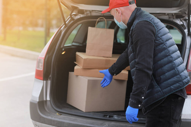 Young delivery man in protective mask, red cap and gloves near the car with boxes and packages, outdoors. Service coronavirus. Online shopping. mock up - Valokuva, kuva
