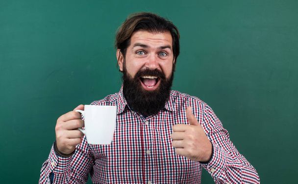 brutal bearded man hold cup of tea and show thumb up. morning energy. just inspired. back to school. formal education. best beverage taste. happy mature teacher drinking morning coffee - Фото, изображение