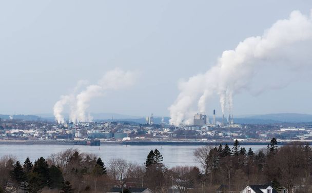 Nehéz ipar a távolban egy víztesten keresztül. Több gyárból is felszálló füst. Kissé ködös a levegő. Kék felhős ég. Az előtérben lévő házak fái és tetejei.  - Fotó, kép