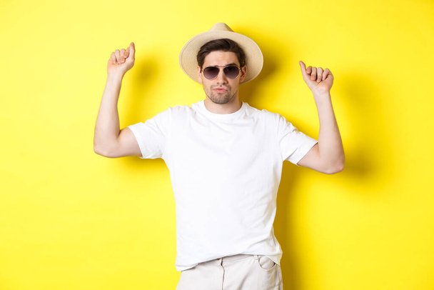 Tourism, travelling and holidays concept. Man tourist enjoying vacation, dancing in straw hat and sunglasses, posing against yellow background - Foto, immagini