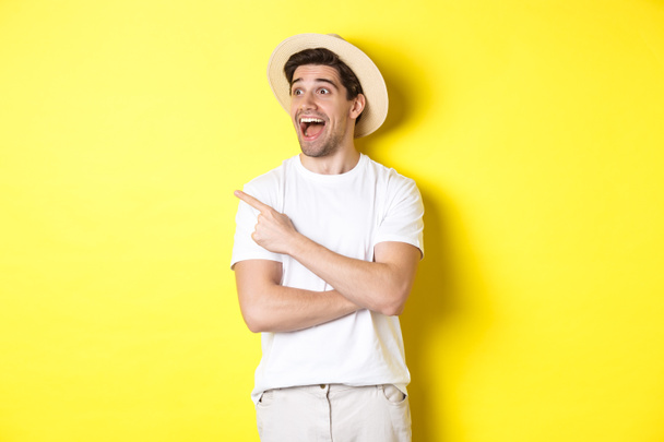 Концепция туризма и образа жизни. Захватывающий красивый парень в соломенной шляпе смотрит рекламу, указывает и смотрит на логотип верхнего левого угла, желтый фон - Фото, изображение