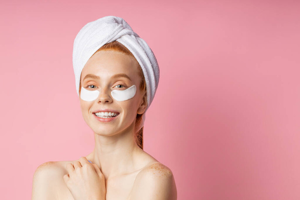 Closeup of beautiful happy caucasian woman with moisturizing under eye mask, towel on head, fresh clean skin, bare shoulders, cute smiling at camera isolated on pink background. Under eye patches. - Photo, Image