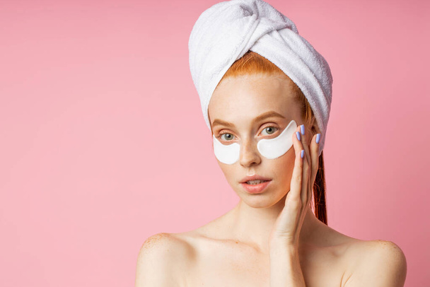 Portrait of beautiful young woman with smooth soft skin applying white under eye hydro gel patches. Charming lady with eye mask, towel on head, keeping palm on cheek posing against pink studio wall. - Fotó, kép