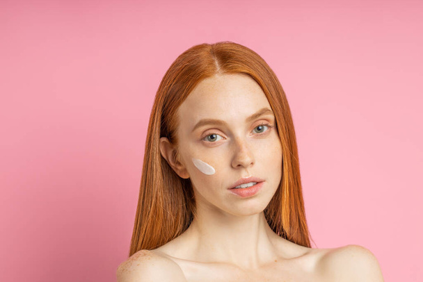 Beauty portrait of young woman with cream on cheek. Cropped shot of lovely naked redhead female using skin care product, moisturizer or lotion, posing on pink background. - Фото, изображение