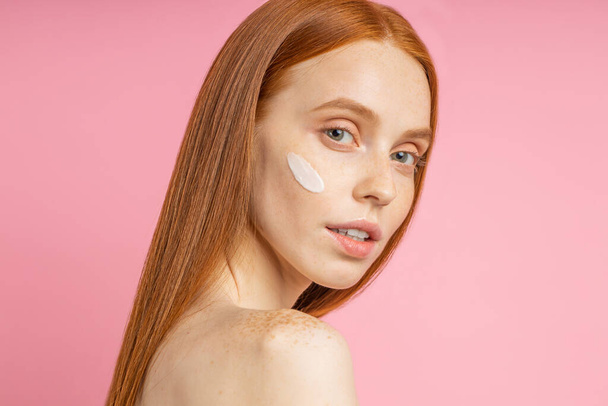 Beauty portrait of young woman with cream on cheek. Cropped shot of lovely naked redhead female using skin care product, moisturizer or lotion, posing on pink background. - Fotoğraf, Görsel