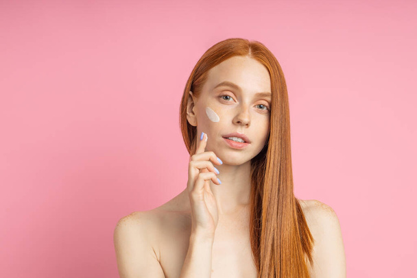 Closeup cropped shot of young caucasian redhead woman applying foundation, face primer or moisturizer on her face. Beauty product, cosmetic, skin care. - Zdjęcie, obraz