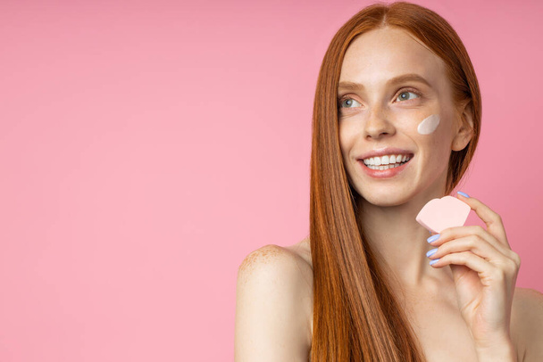 Headshot of beautiful joyful redhead woman with clean healthy skin, applying foundation with cosmetic sponge, smiling showing white teeth isolated on pink. Natural beauty. Skin care. - Photo, Image