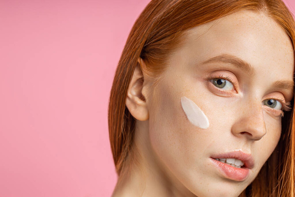 portrait of young woman with cream on cheek. Attractive caucasian woman with red hair, clean fresh freckled skin, posing against pink wall. Natural beauty concept. - Photo, image