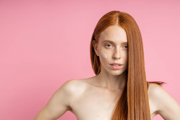 Charming good looking serious girl with perfect freckled skin, naked shoulders, cream on cheek, straightening her long shiny healthy red hair, looking at camera. Skin care, hair treatment, spa. - Фото, зображення