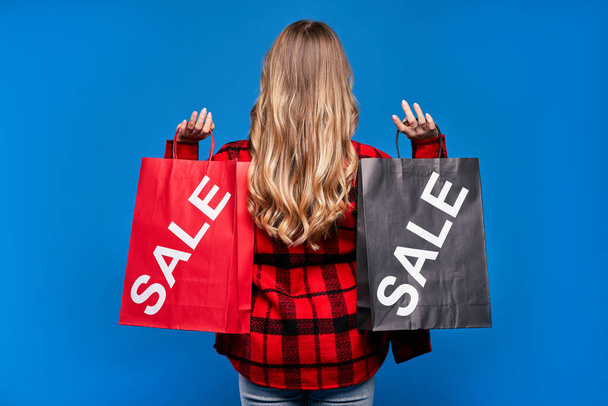 Back portrait young blonde girl in casual clothes on a blue background with a package, autumn sale. - Foto, Imagen
