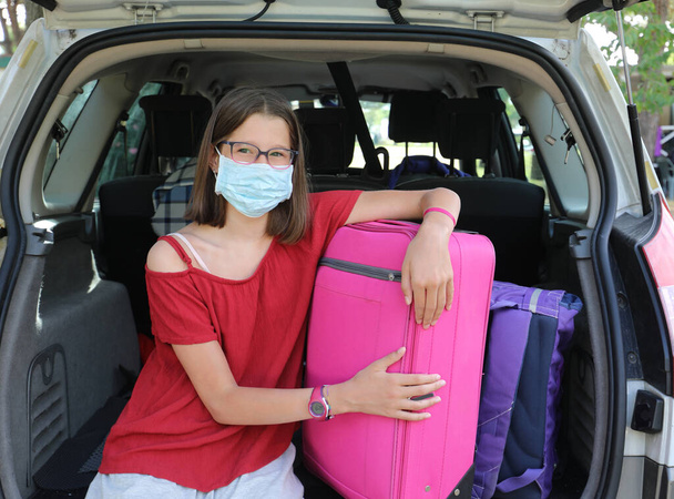 linda niña con máscara quirúrgica para protegerse de coronavirus durante las vacaciones mientras carga el maletero del coche con maletas - Foto, Imagen