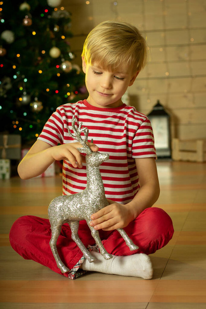 Smiling child in a red Santa Claus hat playing with new year silver deer with on the background of the christmas tree. New Years magic. Christmas concept - Foto, Imagem