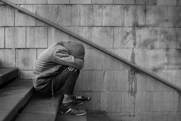 Drunk homeless unhappy man covered his face with his hands and sits on the stairs in the underpass, black and white photo. - Photo, Image