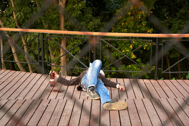 Jeune sans-abri ivre dormant avec une bouteille d'alcool dans les mains sur une surface en bois dans le parc. - Photo, image