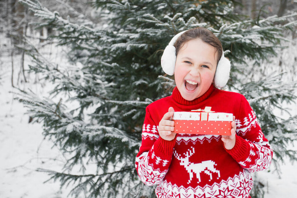 Κορίτσι σε χριστουγεννιάτικο πουλόβερ και ωτοασπίδες με δώρο στο δάσος του χειμώνα - Φωτογραφία, εικόνα