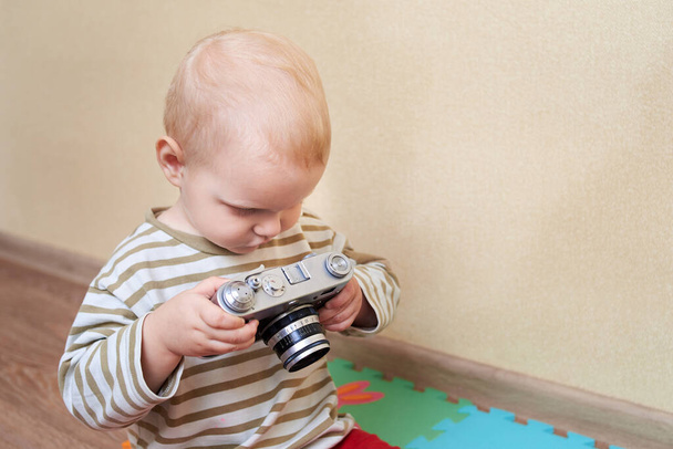 Pequeño chico rubio sostiene una cámara vintage en casa. - Foto, imagen