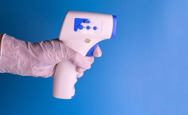 Ανέπαφη θερμόμετρο στο χέρι ενός άνδρα σε ένα ιατρικό γάντι σε ένα γαλάζιο φόντο. Αντιγραφή χώρου. - Φωτογραφία, εικόνα