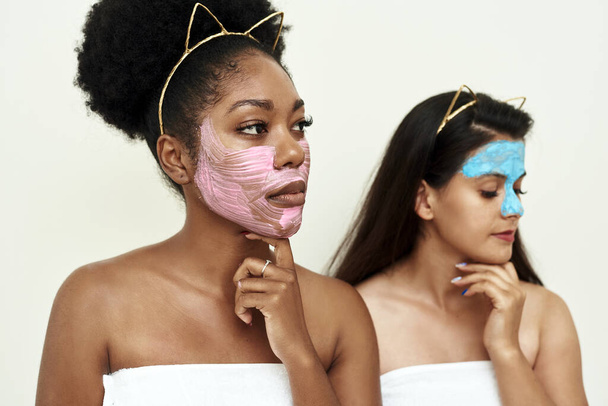 Two girlfriends or young women in love try multimasking on themselves. Spa day. A picture of two girls friends relaxing with facial masks on over white background - Photo, Image