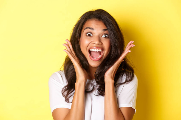Close-up of excited african-american female model, gasping and looking amazed, raising hands up and seing something cool, standing over yellow background - Photo, Image