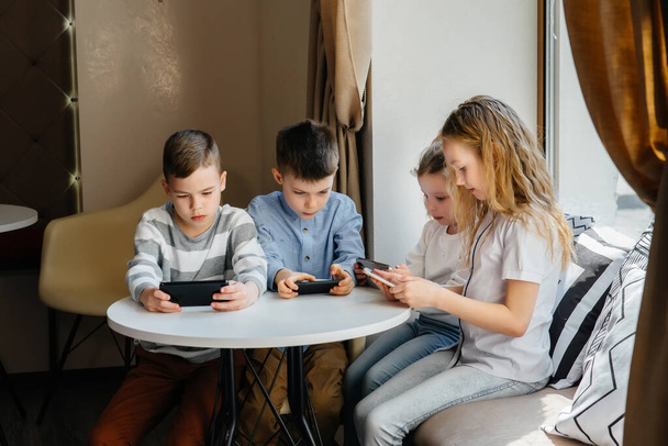 Children sit at a table in a cafe and play mobile phones together. Modern entertainment - Foto, afbeelding