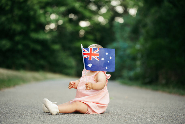 Mignon adorable bébé fille agitant drapeau australien. Enfant assis sur la route dans le parc couvrant visage avec drapeau de la Journée de l'Australie. Célébration de la fête nationale australienne en janvier en plein air. - Photo, image
