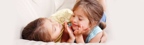 Kaksi söpöä pientä valkoihoista tyttöä, jotka leikkivät kotona. Suloisia hymyileviä lapsia, jotka makaavat sohvalla yhdessä. Aito vilpitön elämäntapa kotielämän hetki. Web-bannerin otsikko. - Valokuva, kuva