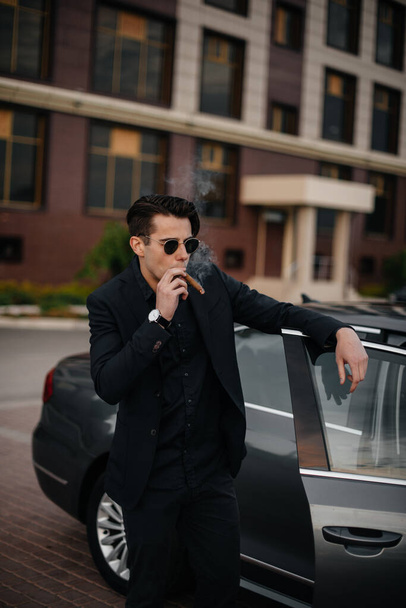 A stylish businessman smokes cigars near a luxury car. Fashion and business - Foto, Bild