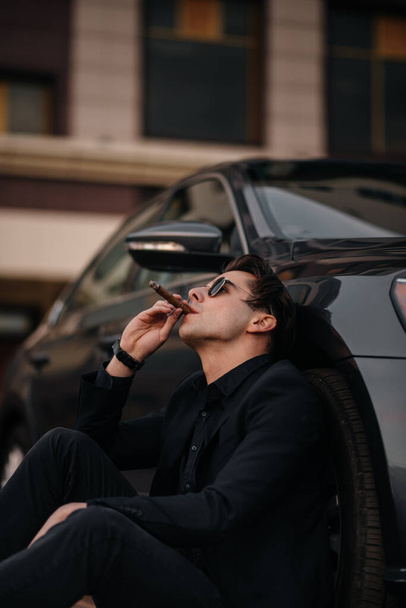 A stylish businessman smokes cigars near a luxury car. Fashion and business - Фото, зображення