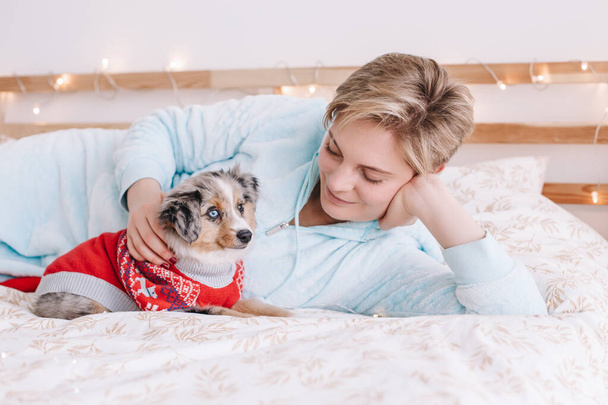 Young Caucasian woman with short hair lying on bed with cute puppy dog at home. Pet owner petting stroking little furry friend. Relationship of domestic animal and human. - Photo, Image