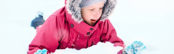 Vtipné běloška dívka v zimním oblečení hrát se sněhem. Roztomilé dítě ležící na zemi během chladného zimního sněhového dne na sněhu. Děti venkovní sezónní aktivity. Záhlaví webového nápisu. - Fotografie, Obrázek