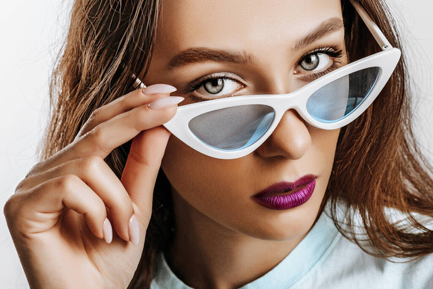 Beautiful young brunette girl with green eyes with fashion makeup with purple lips. The woman in blue sunglasses seriously looking at the camera. Beauty concept. - Fotoğraf, Görsel