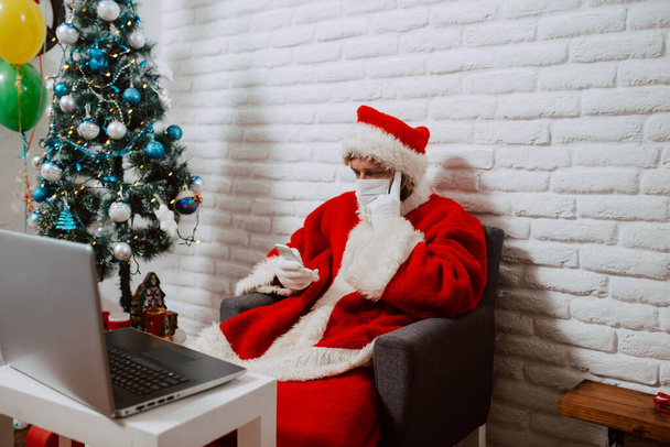 A young Caucasian Santa Claus with a medical face mask sits in the house in front of a laptop and uses the phone. Santa Claus online during COVID - 19 coronavirus - Fotoğraf, Görsel