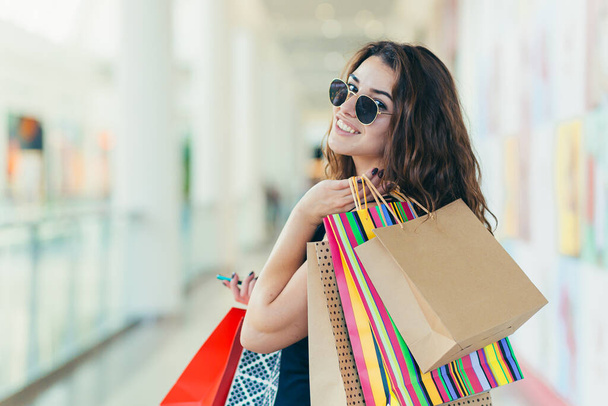 Cheerful lady in stylish black dress carrying on colorful shopping bags while walking at store. Attractive brunette looks satisfied after shopping at mall. - Fotoğraf, Görsel