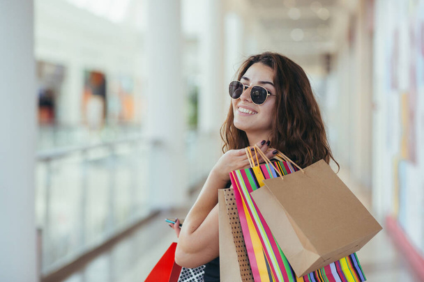Cheerful lady in stylish black dress carrying on colorful shopping bags while walking at store. Attractive brunette looks satisfied after shopping at mall. - Valokuva, kuva