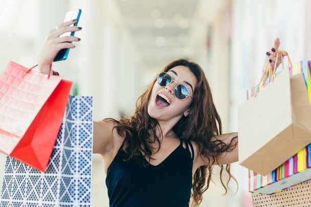 Cheerful lady in stylish black dress carrying on colorful shopping bags while walking at store. Attractive brunette looks satisfied after shopping at mall. - 写真・画像