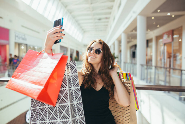 Cheerful lady in stylish black dress carrying on colorful shopping bags while walking at store. Attractive brunette looks satisfied after shopping at mall. - Photo, Image