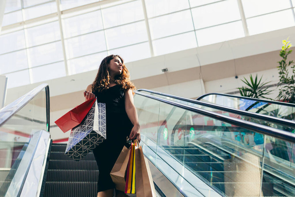 Vrolijke dame in stijlvolle zwarte jurk dragen op kleurrijke boodschappentassen tijdens het lopen in de winkel. Aantrekkelijke brunette kijkt tevreden na het winkelen in winkelcentrum. - Foto, afbeelding