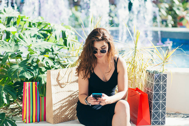 Cheerful lady in stylish black dress carrying on colorful shopping bags while walking at store. Attractive brunette looks satisfied after shopping at mall. - Φωτογραφία, εικόνα