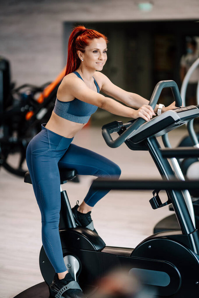 Fit active woman in the gym using gym machines, working out on stationary bicycle.  Fitness and bodybuilding concept - Foto, Imagem