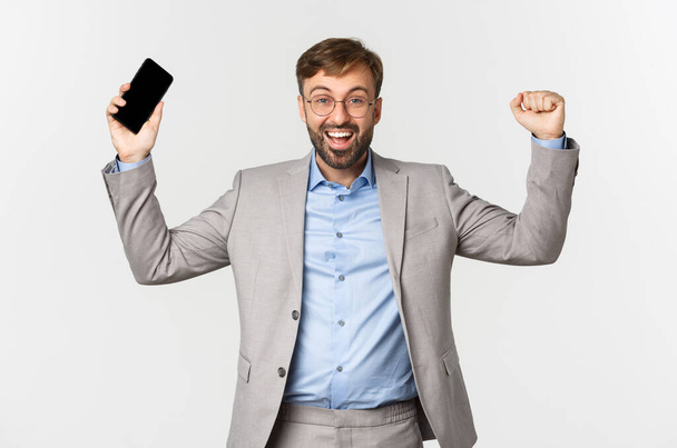 Image of successful businessman in grey suit and glasses, holding mobile phone, raising hands up and shouting yes from satisfaction, triumphing over white background - Foto, Imagen