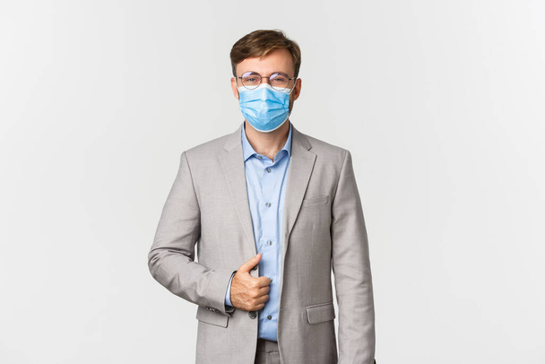 Concept of work, covid-19 and social distancing. Image of confident and successful businessman in gray suit, glasses and medical mask, smiling pleased, standing over white background - Photo, Image