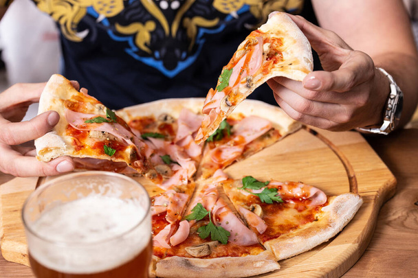 Gustosa pizza con prosciutto e bicchiere di birra sono sul tavolo di legno. Le mani degli uomini tengono un pezzo pizza fresca su una tavola di legno.   - Foto, immagini