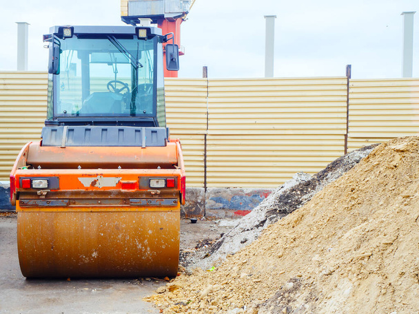 Um rolo para rolar asfalto fica perto de uma pilha de areia no fundo de uma cerca de construção - Foto, Imagem
