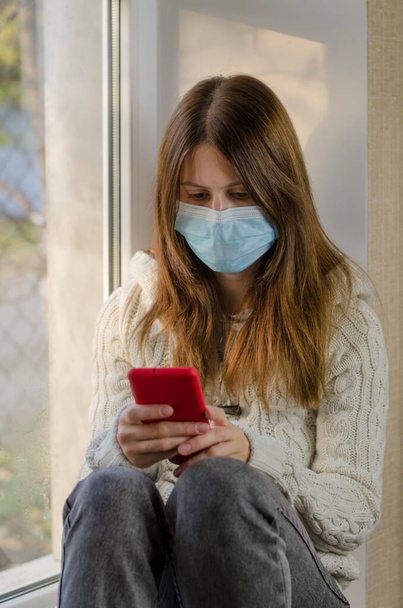 Woman in a medical mask sitting on the windowsill looking into a smartphone - Photo, Image
