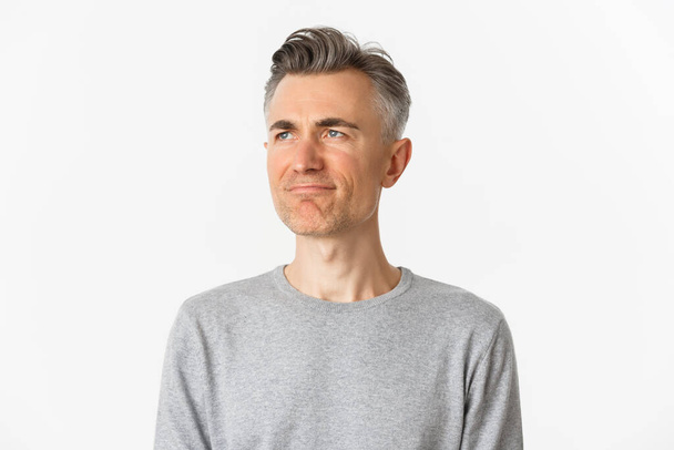 Primer plano del hombre decepcionado de mediana edad en suéter gris, frunciendo el ceño molesto y mirando a la izquierda en el espacio de copia, de pie sobre el fondo blanco - Foto, Imagen