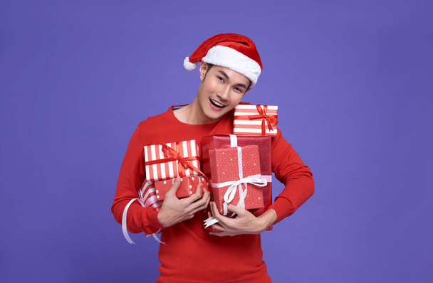 Young asian man in red casual attire wearing Santa hat and holding stack of presents with smile face on purple background.Happy new year concept. - Foto, imagen