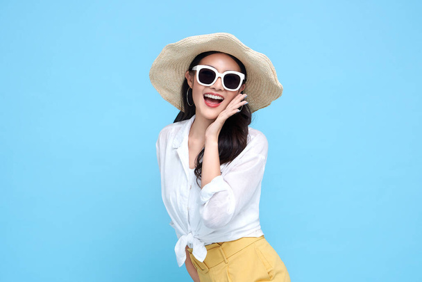 夏のカジュアルな服の若い美しいアジアの女性がわら帽子をかぶって、サングラスは何かを言い、手側の口と笑顔は孤立した明るい青の背景にあります. - 写真・画像